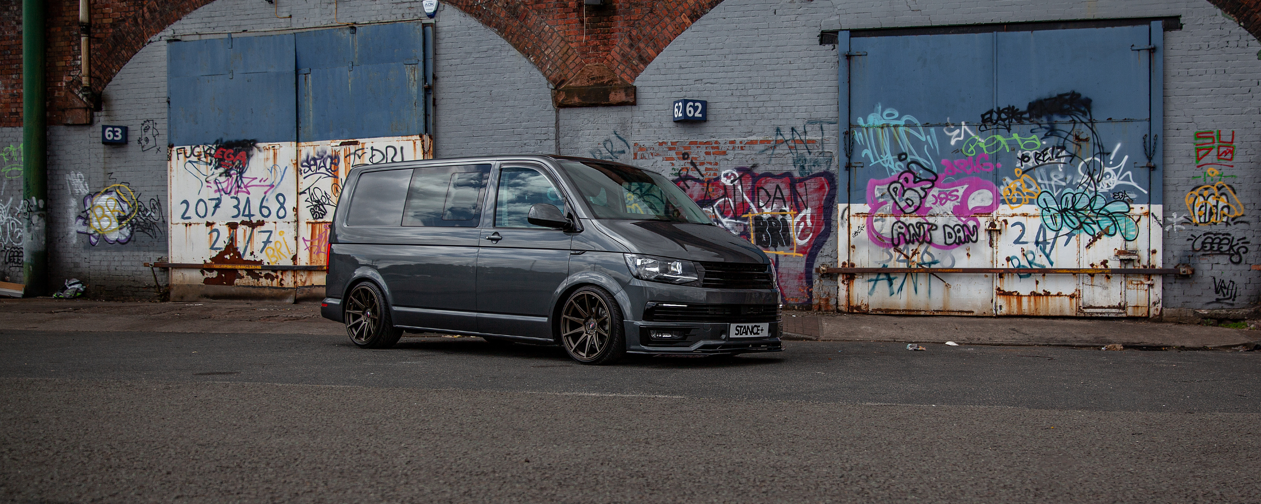 Gray VW Transporter T6 van parked in front of a graffiti-covered wall. Fitted with Stance+ Street Coilover Suspension.